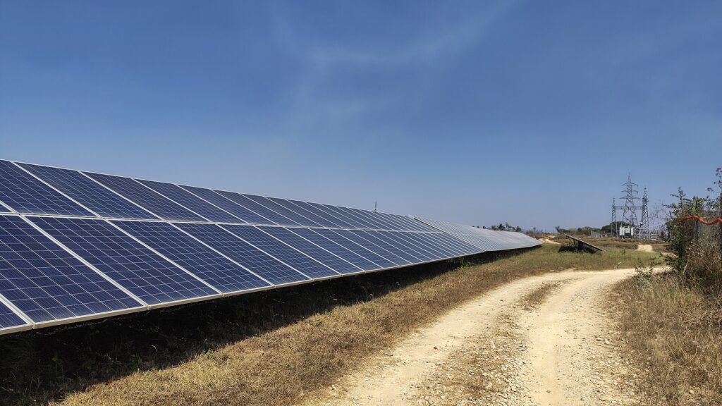 Row of solar panels beside a dirt track with a grid transmission tower in the background in India